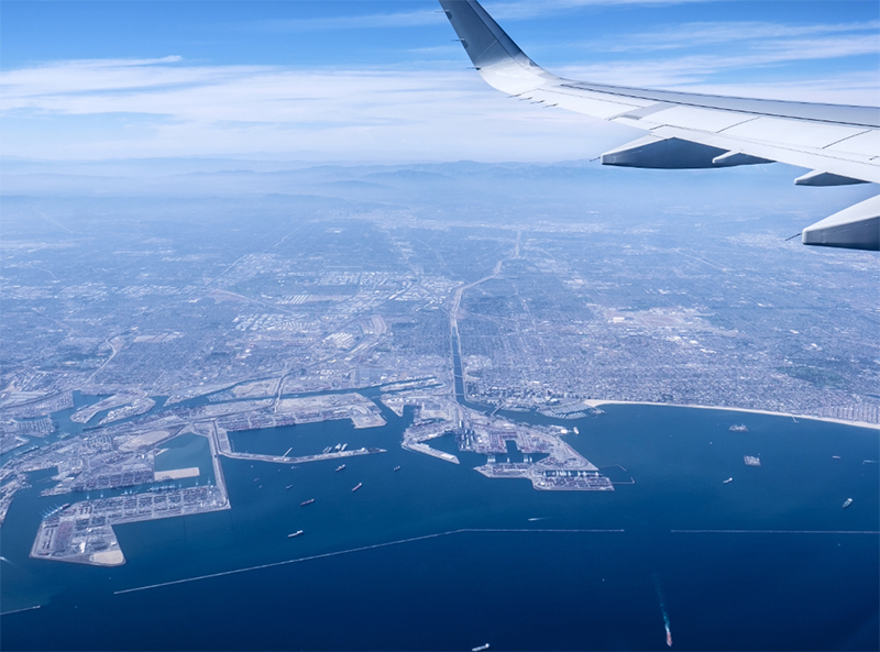 Aerial view from an airplane over the Port of Los Angeles and Long Beach, showing the coastline, harbor infrastructure, city, and aircraft wing. Marketing Launch Kit Case Studies.